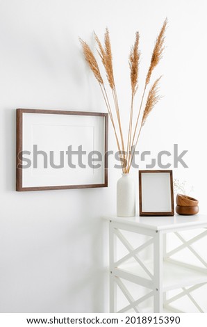 Stylish frames mockup for artwork, print or photo presentation. Blank wooden frames on white wall with furniture and ceramic vase with dry plant decorations.