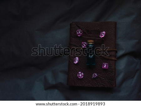 Image of pink transparent gaming dice and a glass vial with dark liquid on a leather-bound notebook in the shadow