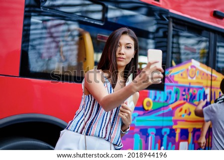 Youthful Asian female tourist making selfie pictures for creating travel content for web blog using front camera on modern mobile technology, charming hipster girl shooting influence video