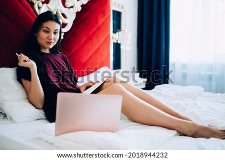 Millennial female IT professional watching education webinar while planning organisation of distance job, skilled Asian freelancer with textbook using laptop computer and WIFI interner in hotel room