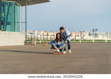 Young caucasian couple enjoy skating together on street. Hipster man pushing back of woman sitting on longboard. Cheerful lovers or friends have fun outdoors over city skyline. Urban lifestyle concept
