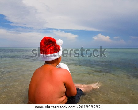 a man in the image of Santa Claus in a festive hat is sitting on the beach and looking at the sea. vacation by the ocean at Christmas. new Year holidays in warm countries