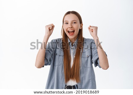 Excited teenage girl rejoicing, shouting yes with joy and fist pump, celebrates victory, achieve goal, wins and triumphs, becomes champion, standing over white background