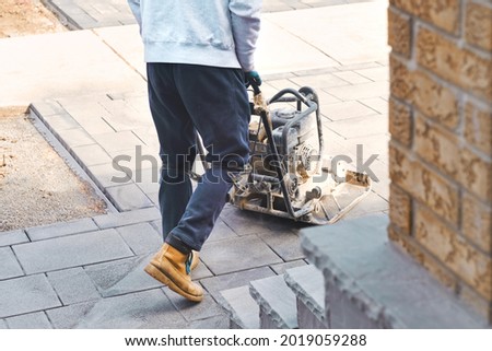Landscaping worker contractor using plate compactor to apply polymeric joint sands on interlock pavers. Home garden and landscape renovation project background.  Royalty-Free Stock Photo #2019059288