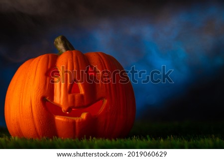 An interesting original composition for the Halloween holiday. Against the background of a dark cloudy sky with haze on the green grass, a large orange pumpkin with a creepy smile. Close-up.