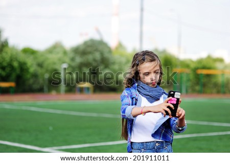 Adorable pre-teen tweenie kid girl making self portrait with funny duck face on her cell phone on the school soccer field