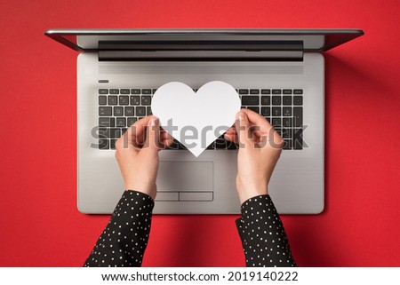 Overhead photo of grey laptop and hands holding a white paper heart isolated on the red backdrop