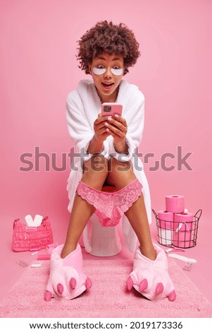 Surprised curly haired woman sits comfortably on toilet bowl uses mobile phone for chatting online dressed in white bathrobe poses in restroomm applies beauty pads under eyes isolated on pink wall
