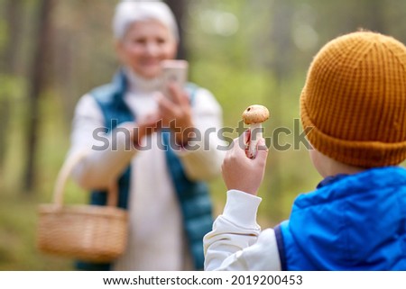 picking season, leisure and people concept - close up of grandmother with smartphone photographing happy smiling grandson with mushroom in forest