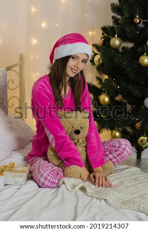 Christmas, beautiful woman in pajamas and a santa claus hat in a new year's interior on a bed with a toy bear.