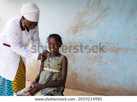 Black pediatrician with a doctor's suit is placing a stethoscope on the chest of a smiling little girl with typical African braids during routine respiratory check-up outdoors a rural hospital setting Royalty-Free Stock Photo #2019214985