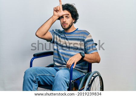 Handsome hispanic man sitting on wheelchair making fun of people with fingers on forehead doing loser gesture mocking and insulting. 