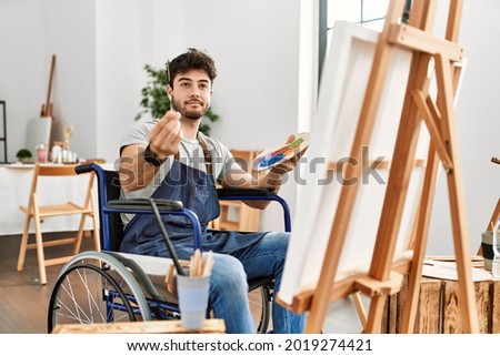 Young hispanic man sitting on wheelchair painting at art studio doing money gesture with hands, asking for salary payment, millionaire business 