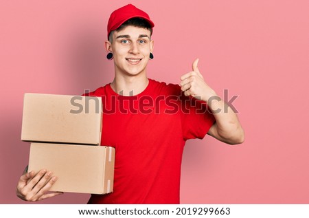 Young caucasian boy with ears dilation holding delivery package smiling happy and positive, thumb up doing excellent and approval sign 