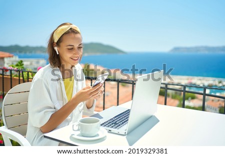 Young elegant woman wearing wireless headphone looking at smartphone with smile while sitting at balcony with seaview on resort. girl watching video, chatting in social network Royalty-Free Stock Photo #2019329381