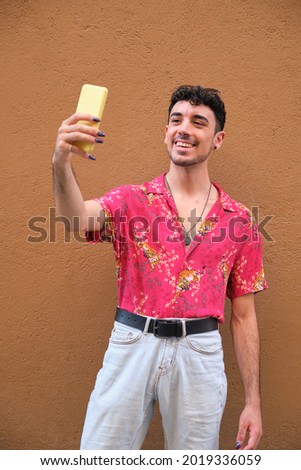 Young caucasian man with long false nails taking a selfie with his smartphoneon a brown wall.