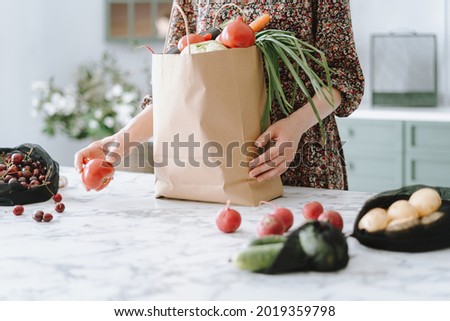 Woman in dress unpacking vegetables from paper grocery bag onto kitchen island with marble top, getting ready to prepare healthy and nutritious meal, furniture in blurred background, cropped shot Royalty-Free Stock Photo #2019359798