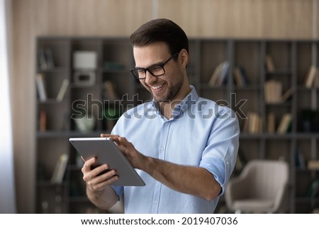 Smart tech. Positive guy in spectacles use touch pad device to learn or work online choose goods services at web shop scroll news at social media. Young man stand at living room play game on tablet pc Royalty-Free Stock Photo #2019407036