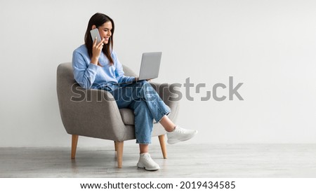 Full length of Caucasian lady using laptop and speaking on smartphone while sitting in armchair against white studio wall, free space. Lovely young woman having phone conversation, working on pc Royalty-Free Stock Photo #2019434585