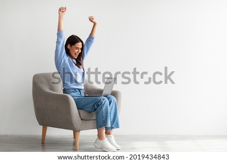 Overjoyed millennial woman sitting in armchair with laptop, celebrating huge online win or success against white studio wall, copy space. Young lady enjoying big sale in web store