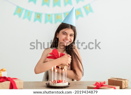 Happy Indian teen girl in party hat sitting at table, hugging gift box, having birthday cake with candles at home. Cheerful Asian adolescent celebrating holiday alone, looking at camera Royalty-Free Stock Photo #2019434879