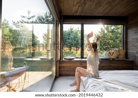 Woman wakes up in a country house or hotel with panoramic windows in pine forest raised her hands yawning. Good morning and recreation on nature concept Royalty-Free Stock Photo #2019446432