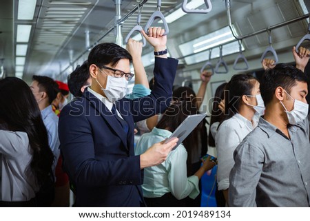 Asian man wearing medical face mask reading news or checking work on computer tablet in subway with crowded people. new normal lifestyle during coronavirus pandemic Royalty-Free Stock Photo #2019489134