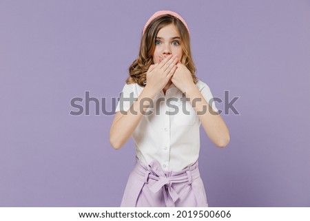 Little shocked surprised blonde kid girl 12-13 years old in white short sleeve shirt covering mouth with hands isolated on purple color background children studio portrait. Childhood lifestyle concept