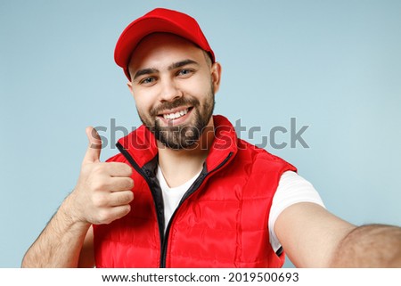Close up professional delivery guy employee man in red cap white T-shirt uniform workwear work as dealer courier doing selfie isolated on pastel blue color background studio portrait. Service concept