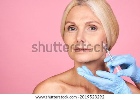 Portrait of calm serious charming aged woman with wrinkle getting injection in cheek in professional clinic hands in gloves making face contouring. Isolated on pink background.