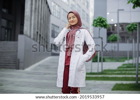 Outdoor portrait of beautiful Malay Muslim nurse with stethoscope. Confident Arab female doctor in traditional headscarf standing outside modern clinic or hospital buildings