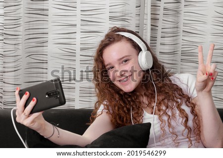 Caucasian girl with smartphone in hand and headphones, smiles at the camera and makes peace sign sitting in her living room.