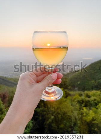 Rising sun in glass of white wine in an outstretched hand against the backdrop of green valley, morning sky. Summer time. Concept of relaxing vacation, travel, wine tasting, romance. Georgia. Vertical