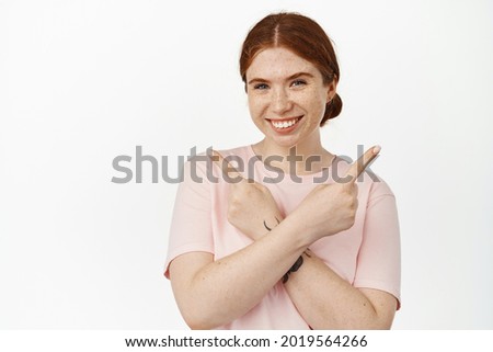 Image of smiling ginger girl pointing sideways with crossed arms, showing left and right product deals, store offers, looking happy, standing against white background