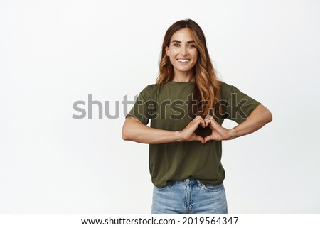 Image of adult woman showing heart sign, I love you gesture, smiling happy and caring, express sympathy, like someone, standing over white background