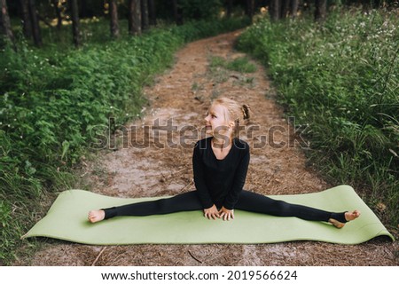A beautiful little girl, a child, a professional gymnast in a black suit trains diligently, stretches in a twine pose in the woods in nature on a green long rug.