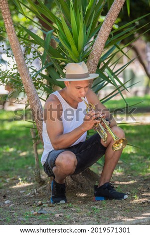 Latino man playing the trumpet crouched near a plant outdoors