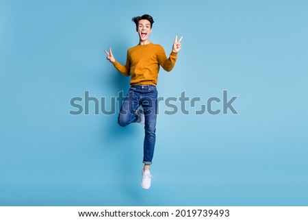 Full length photo of cool brunet young guy jump show v-sign wear sweater jeans isolated on blue color background
