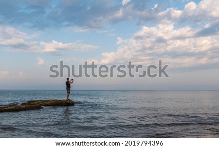 Young caucasian man in shorts taking photo with mobile phone on Croatia sea rock shore edge under cloudy sky. Side view, copy space