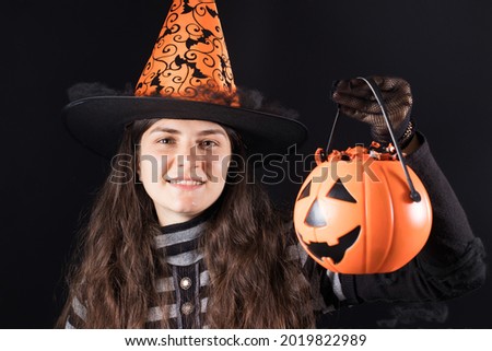 A woman in a witch costume in a hat and gloves on a black background holds bucket pumpkin with candy and smile. Halloween people