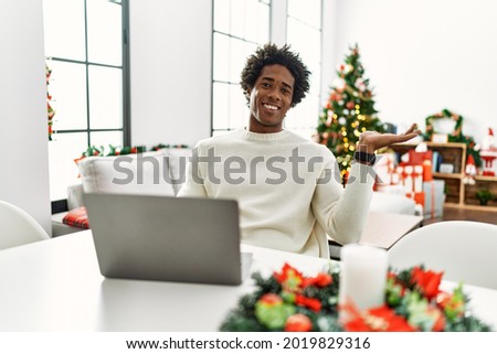 Young african american man using laptop sitting on the table by christmas tree smiling cheerful presenting and pointing with palm of hand looking at the camera. 