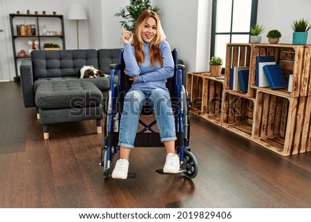 Young beautiful woman sitting on wheelchair at home with a big smile on face, pointing with hand and finger to the side looking at the camera. 
