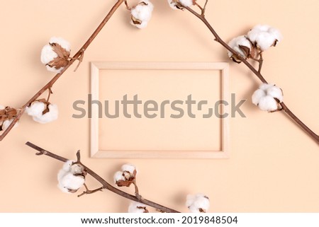 Empty wooden frame and cotton branches on a beige background. Creative concept with copy space.