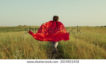 Child plays and dreams. Happy superhero girl, runs on green field in red cloak, cloak flutters in wind. Teenager dreams of becoming superhero. Young girl in red cloak, dream expression. Royalty-Free Stock Photo #2019852458