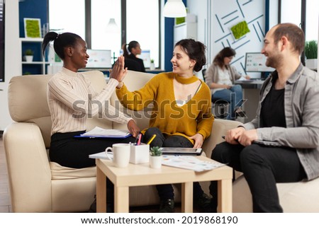 Confident company manager giving working tasks to diverse teammates sitting on couch in start up office. Multiethnic team discussing project ideas at brainstorming meeting using digital devices