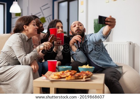 Diverrse group of coworkers taking selfie on smartphone after work at office party. Cheerful colleagues having fun at celebration meting with pizza chips bottles and cups of beer alcohol