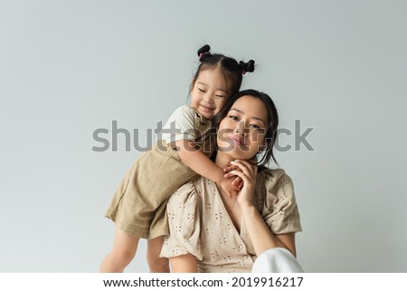 cheerful asian toddler daughter hugging happy mother isolated on gray Royalty-Free Stock Photo #2019916217