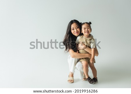 full length of stylish asian mother hugging toddler daughter on grey