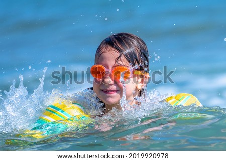 Young beautiful girl having fun in sea surf on beach. Joyful child swimming on wave. Travel, lifestyle, swimming activities and  summer holiday concept. 