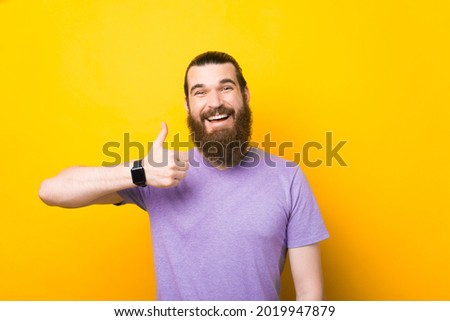 Man with beard wearing purple tshirt standing doing happy thumbs up gesture with hand. Approving expression looking at the camera
